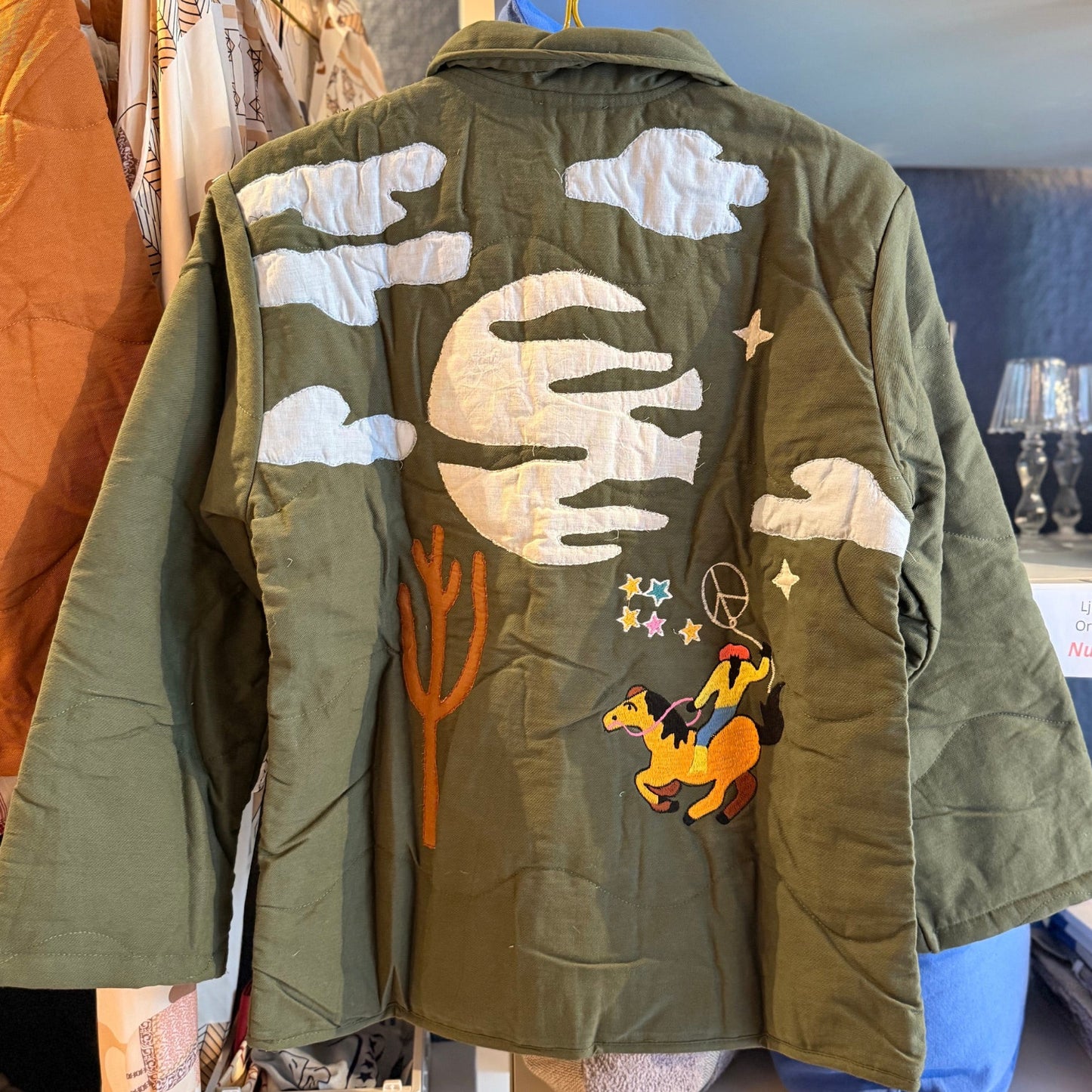 Quiltad jacka med westernmönster med broderad cowboy, häst, kaktusar och moln.
Allt gjort i en cool lappteknik. Här har du en nättare jacka med skjortkrage, knäppning och två fickor. Vi älskar den! The Corner, Paris. Sommarjacka.